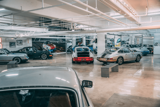 A well-lit underground garage displaying a collection of classic sports cars, mostly Porsches. The cars are arranged in a spacious area with polished concrete floors and painted walls featuring car-related artwork.